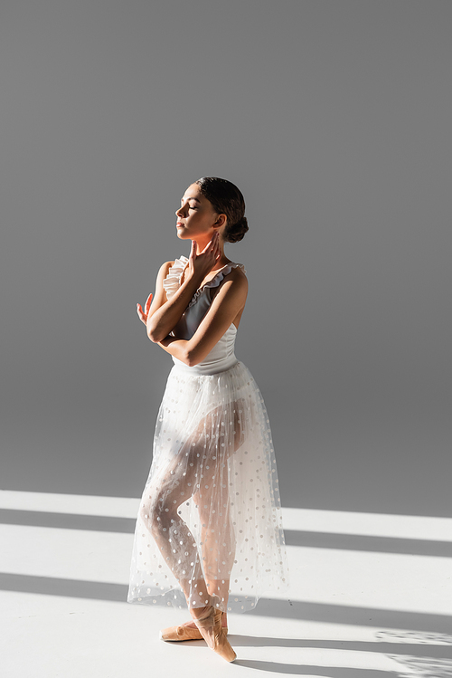 Side view of ballerina with closed eye touching neck on grey background with sunlight