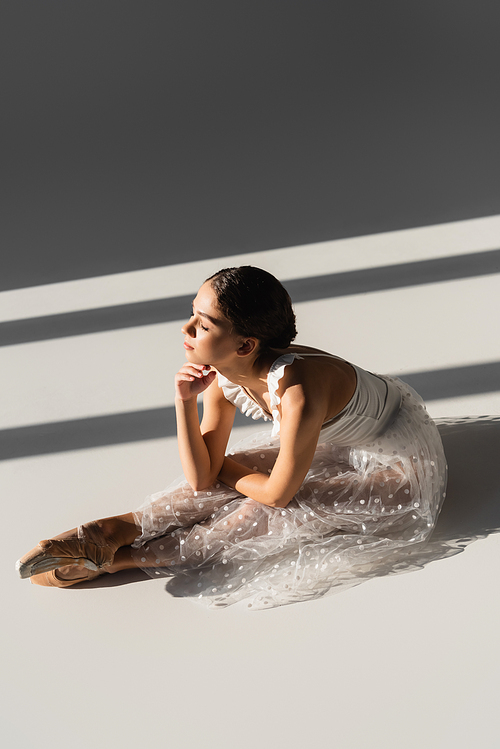 Side view of ballerina bending while sitting on grey background with sunlight