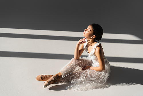 Side view of ballerina with closed eyes sitting on grey background with sunlight