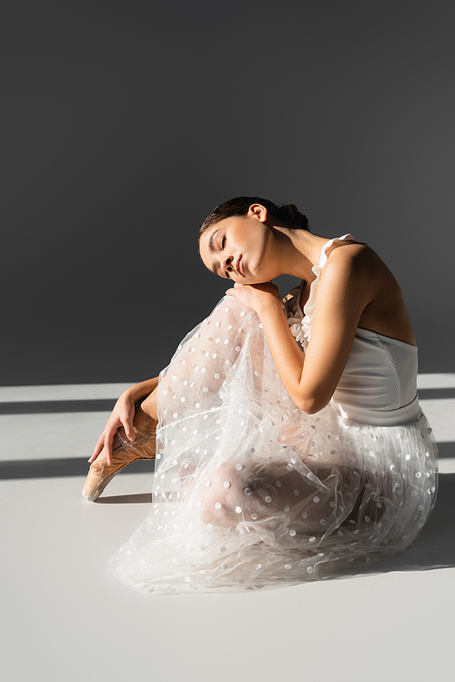 Young ballerina in while dress and pointe shoe sitting on grey background with sunlight