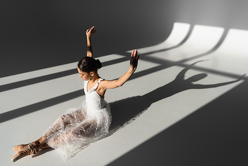 Side view of ballerina raising hands while sitting on grey background with sunlight