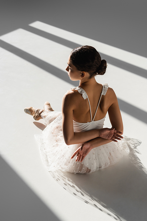 Side view of ballerina in white dress and pointe shoes sitting in sunlight on grey background