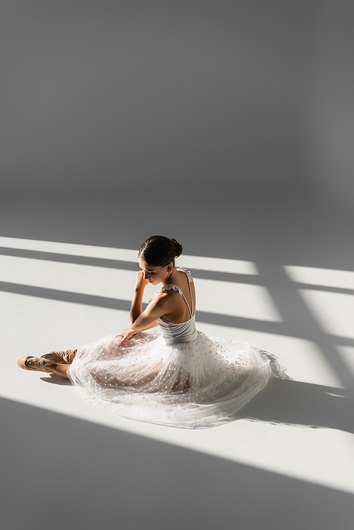 Pretty ballerina in while skirt sitting on grey background with sunlight and shadow