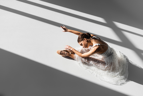 High angle view of pretty classical dancer bending on floor on grey background with sunlight