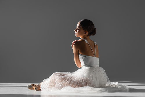 Side view of young ballerina touching shoulder on grey background with sunlight