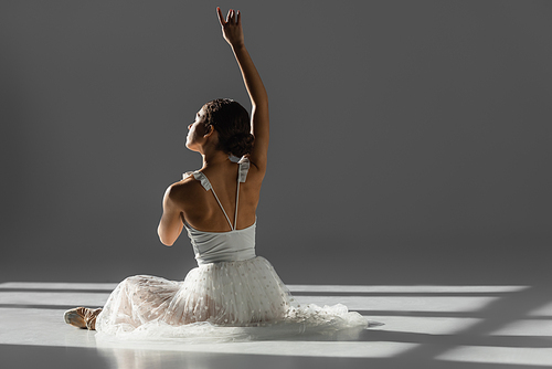 Ballerina raising hand while sitting in sunlight on grey background with sunlight