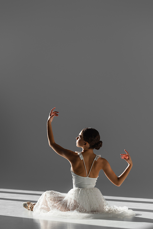 Side view of ballerina in white dress and pointe shoe dancing on floor on grey background with sunlight