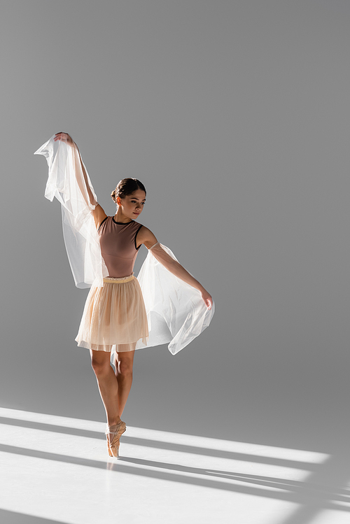 Pretty ballerina in beige skirt dancing with cloth on grey background with sunlight