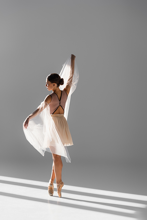 Side view of elegant ballerina dancing with white cloth on grey background with sunlight