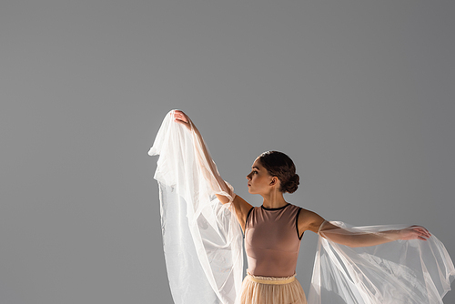 Side view of brunette ballerina dancing with fabric isolated on grey