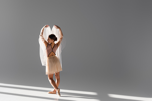 Side view of ballerina with cloth raising hands on grey background with sunlight