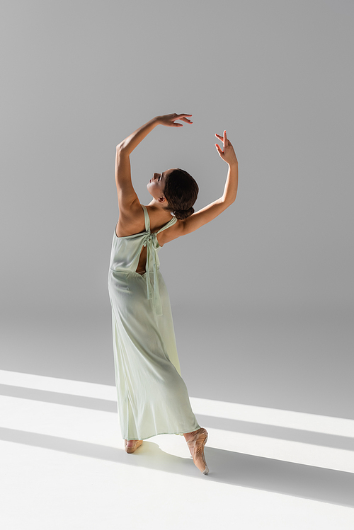 Side view of young dancer in pointe shoes standing on grey background with sunlight