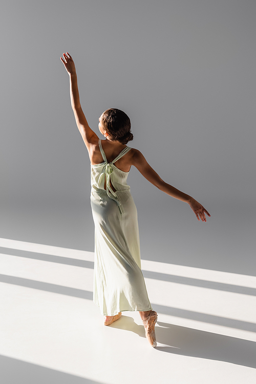 Back view of ballerina in dress raising hand on grey background with sunlight