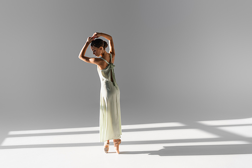 Side view of brunette ballerina in pointe shoes and dress on grey background with sunlight