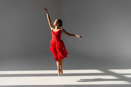 Side view of ballerina in red dress jumping on grey background with sunlight