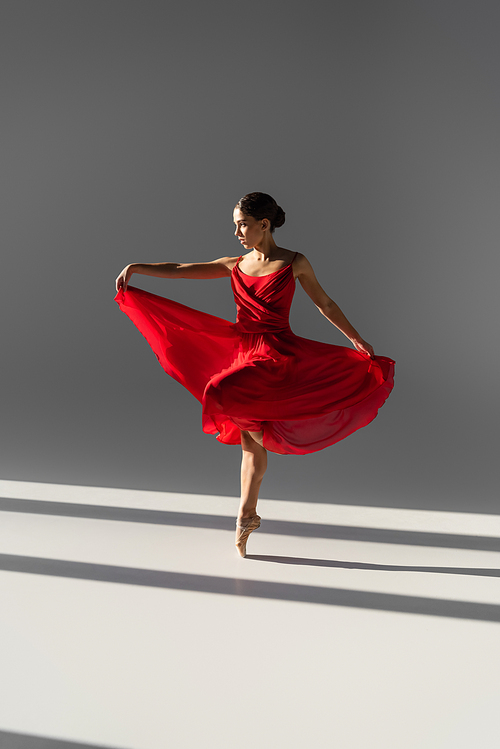 Side view of ballerina dancing and holding red dress on grey background with sunlight