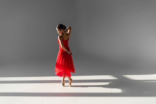 Side view of ballerina in red dress and pointe shoes on grey background with sunlight