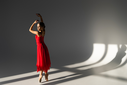 Pretty ballerina in red dress dancing in sunlight on grey background