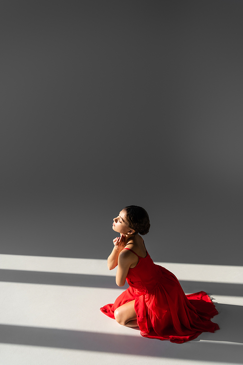 Side view of brunette dancer in red dress sitting on grey background with sunlight