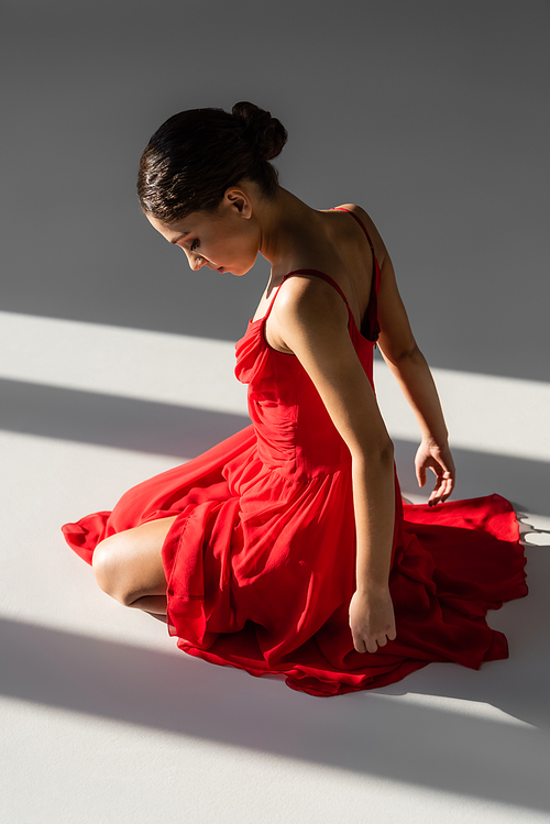 Side view of brunette ballerina in red dress sitting in light on grey background