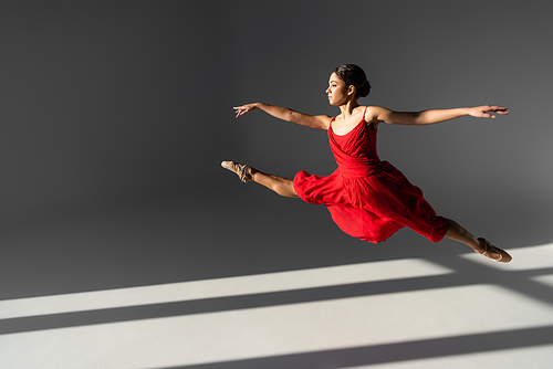 Side view of ballerina in red dress jumping on grey background with sunlight