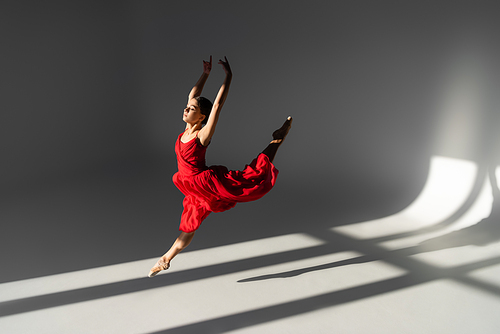 Side view of pretty ballerina in red dress jumping and raising hands on grey background with sunlight