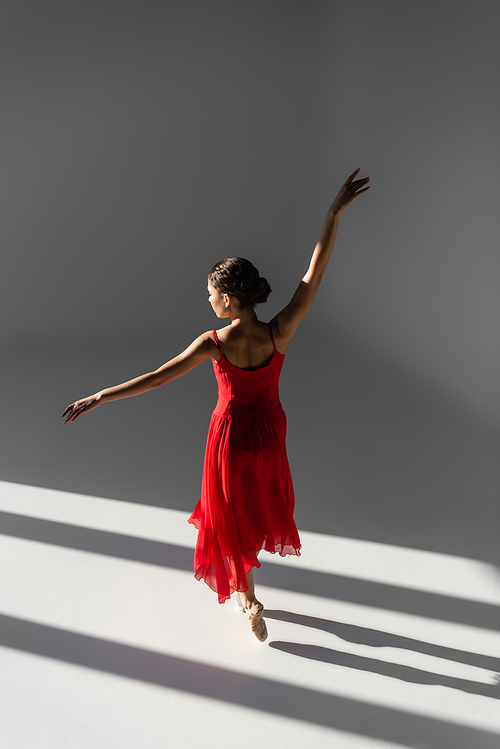 Ballerina in pointe shoes and red dress standing on grey background with sunlight