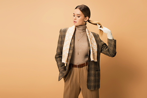 stylish woman in checkered blazer, pants and white gloves posing with hand in pocket on beige