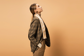side view of stylish woman in checkered blazer posing with hand in pocket on beige