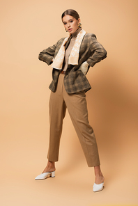 full length view of elegant woman in blazer and pants posing with hands on hips on beige
