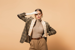 trendy woman in blazer and pants touching forehead while posing with hand on hip on beige