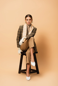 elegant woman in trousers, blazer, white shoes and gloves sitting on beige background