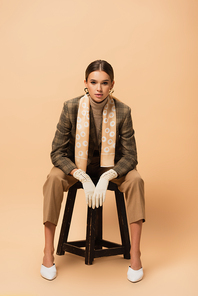 front view of trendy woman sitting on stool in blazer, pants and white gloves on beige