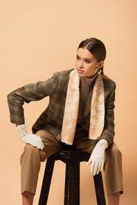 fashionable woman in blazer, pants and gloves looking away while sitting on stool isolated on beige