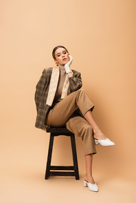 fashionable woman in blazer and pants sitting on wooden stool on beige background