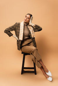 fashionable woman in brown blazer and trousers touching hair while sitting on beige