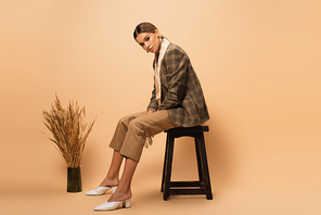 young woman in trendy blazer and pants sitting on stool near vase with spikelets on beige