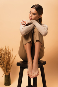 young woman in trendy pants and turtleneck sitting on chair barefoot on beige background