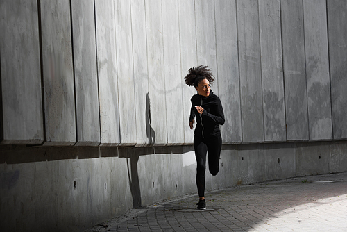 Young african american woman running while training near building with concrete wall