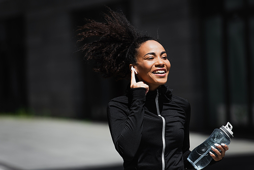 Positive african american jogger with sports bottle using earphone outdoors
