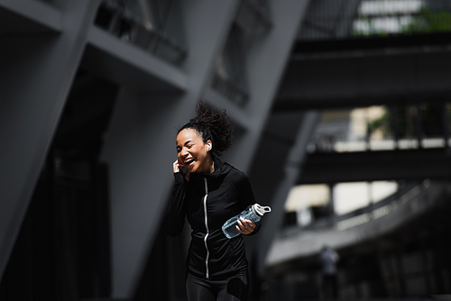 Happy african american sportswoman in earphone holding sports bottle on urban street