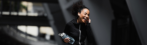 Cheerful african american woman in sportswear listening music in earphone and holding sports bottle outdoors, banner