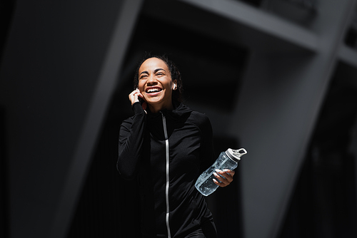 Smiling african american sportswoman in earphones holding sports bottle outdoors