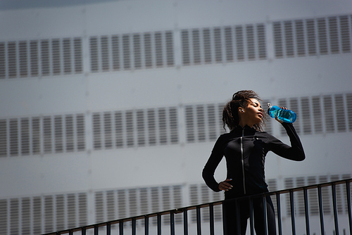 Low angle view of african american sportswoman in earphone drinking water on urban street