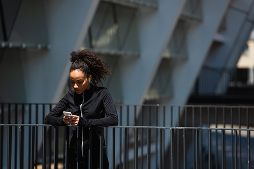 African american sportswoman in earphone using smartphone on urban street