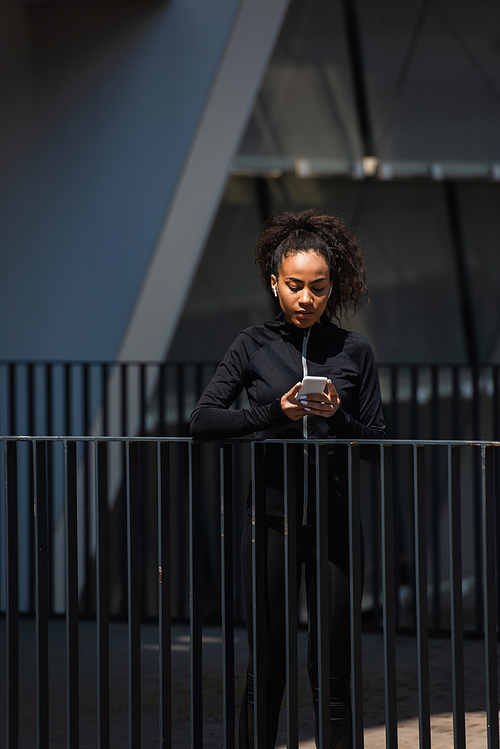 African american sportswoman in earphones using smartphone outdoors