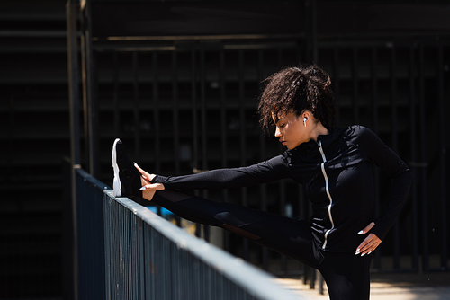 Side view of african american sportswoman in wireless earphone stretching near bridge on urban street