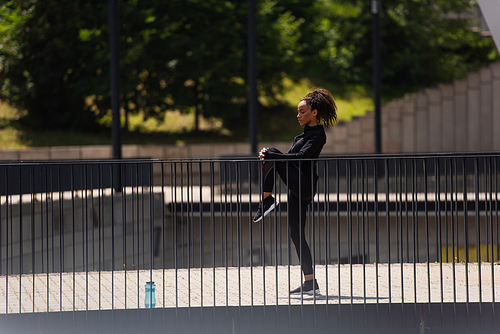Side view of fit african american sportswoman stretching legs near sports bottle on bridge