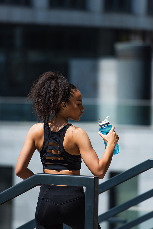 Side view of pretty african american sportswoman holding sports bottle with water on urban street