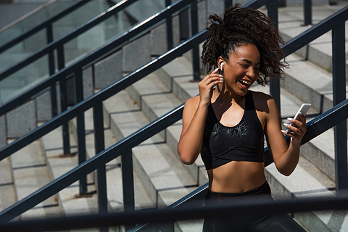 Smiling african american sportswoman holding smartphone and listening music on stairs